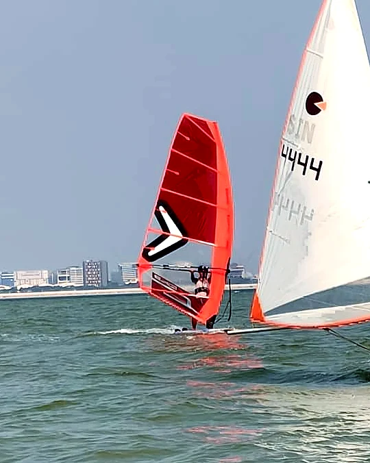 A windsurfer with a red sail alongside a SeaQuest dinghy at Forest City